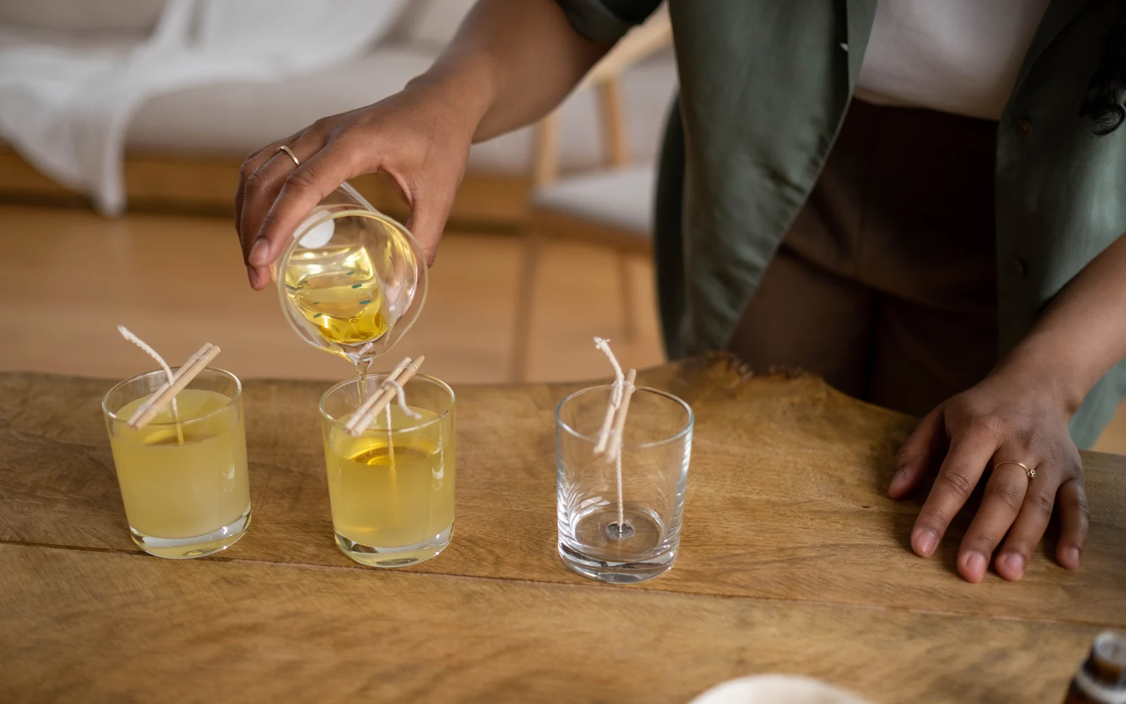 Person pouring liquid wax into multiple clear candle vessels with centered wicks